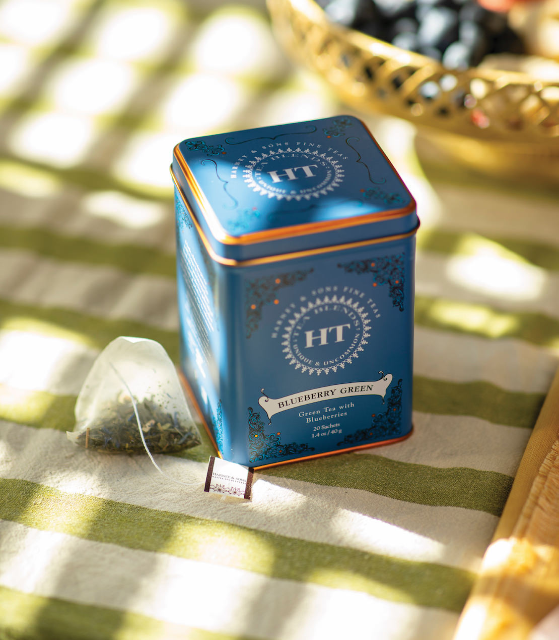 Blueberry Green tea tin with 20 sachets, featuring a vibrant blue design and a sachet beside it on a striped tablecloth.