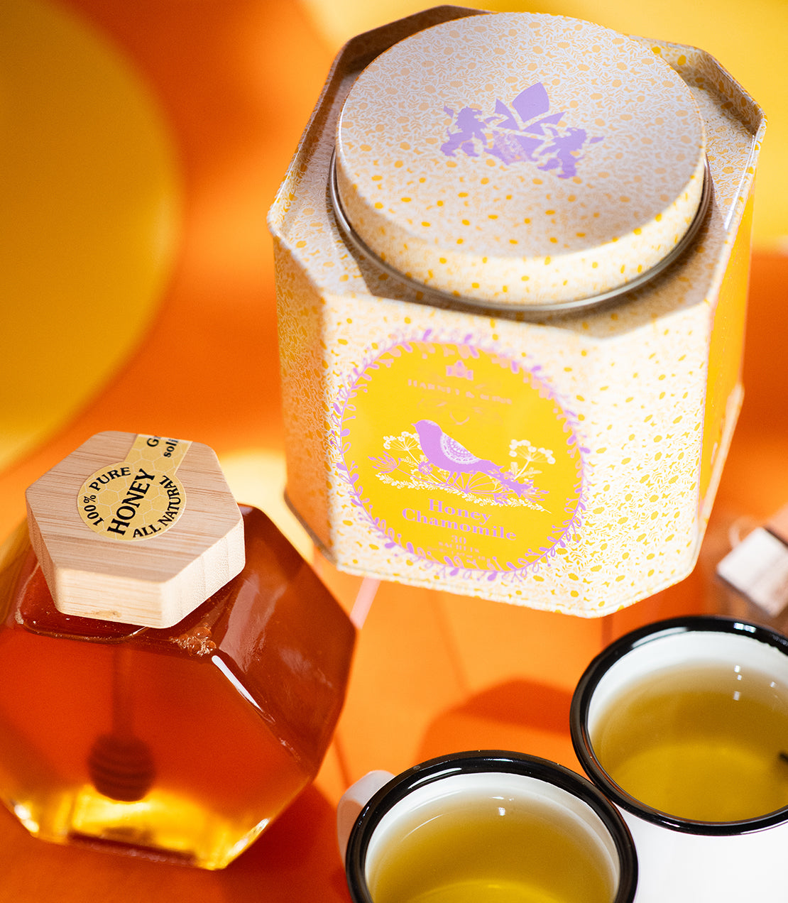 Honey Chamomile tea sachets in a decorative tin, accompanied by a honey jar and two cups of brewed tea.