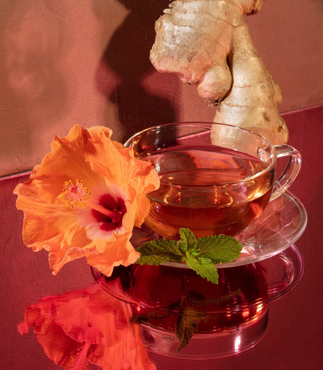 A clear cup of organic hibiscus ginger tea with a vibrant hibiscus flower and fresh ginger root on a reflective surface.