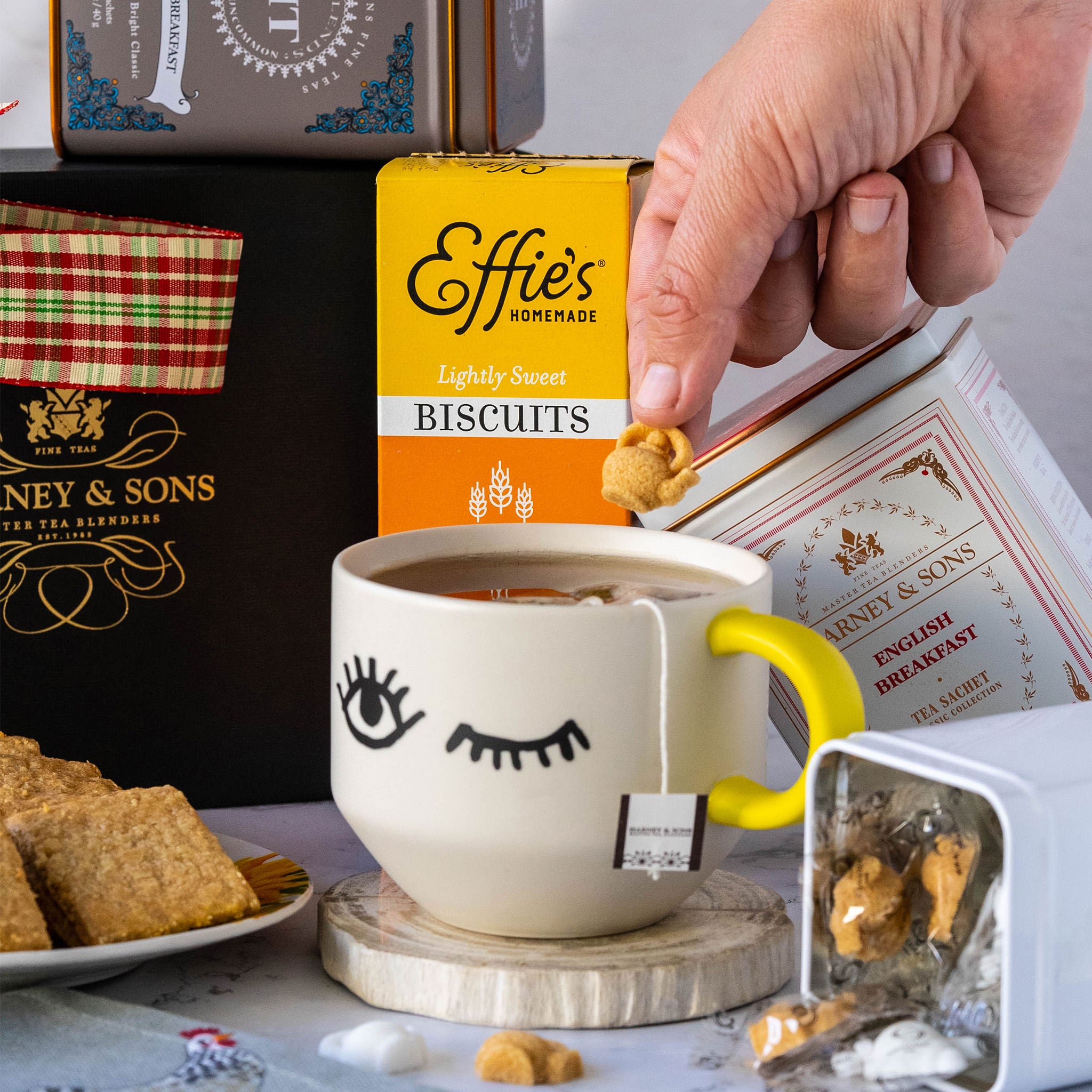 Hand dipping a biscuit into a cup of tea, surrounded by English Delight gift items including tea and snacks.